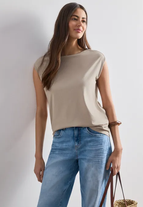 Frau in ärmellosem beigefarbenem Top und blauen Jeans hält eine braune Tasche.