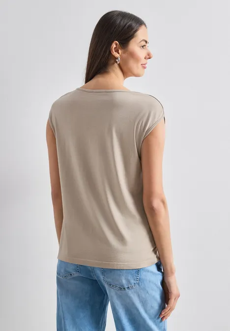 Eine Frau mit langen dunklen Haaren steht von hinten, trägt ein beiges Top und blaue Jeans vor hellem Hintergrund.
