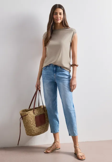 Frau in beigem Top, blauen Jeans, Sandalen und großer Korbtasche vor neutralem Hintergrund.
