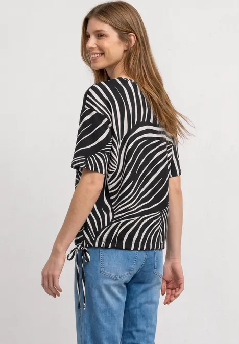 T-shirt à col rond et cordon de serrage Black Femme portant un top noir à imprimé zèbre blanc, manches mi-longues et cordons sur le côté, avec jean bleu.