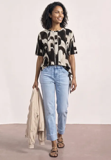 Frau in gemusterter Bluse, hellblauen Jeans und Sandalen hält eine Jacke in der Hand.