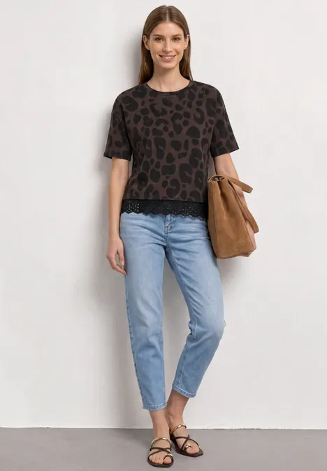 T-Shirt mit Rundhals und Spitze molten earth Frau in Leo-T-Shirt, hellblauen Jeans, schwarzen Sandalen und brauner Tasche vor neutraler Wand.