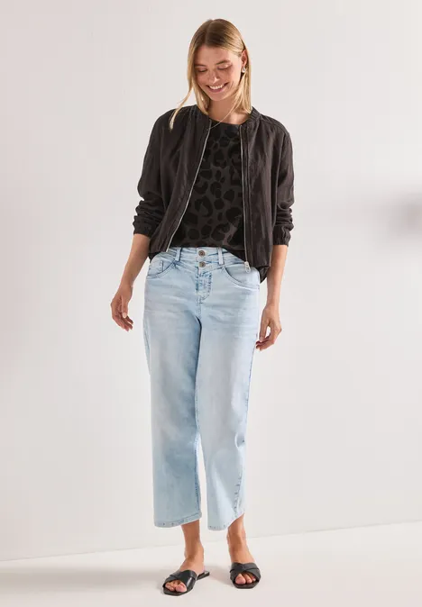 Frau mit schwarzer Bomberjacke, hellblauen Jeans und Sandalen vor weißem Hintergrund