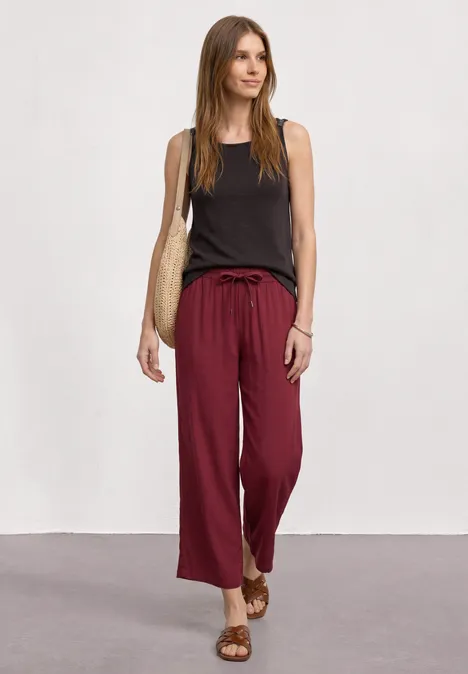 Top mit U-Boot-Ausschnitt und Schulterdetail espresso brown Frau trägt schwarzes Top, weite bordeauxrote Hose, Sandalen und Korbtasche vor neutralem Hintergrund.