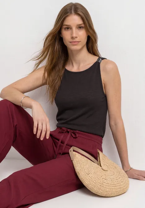 Top mit U-Boot-Ausschnitt und Schulterdetail espresso brown Frau mit schwarzem Top und weinroten Hosen sitzt vor weißem Hintergrund, Strohtasche liegt neben ihr