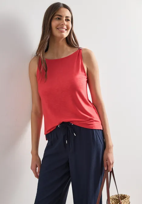 Top mit U-Boot-Ausschnitt und Schulterdetail sunset red Frau in rotem Top und dunkelblauer Hose lehnt lässig an der Wand mit Tasche.