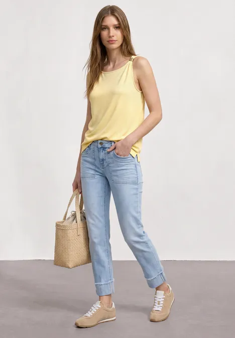 Top mit U-Boot-Ausschnitt und Schulterdetail soft yellow Frau trägt gelbes Top, blaue Jeans, beige Sneaker und Strohtasche vor neutralem Hintergrund.