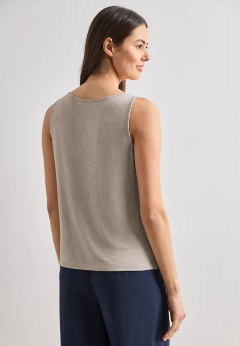 Eine Frau steht von hinten, trägt ein ärmelloses graubraunes Top und dunkelblaue Hose.