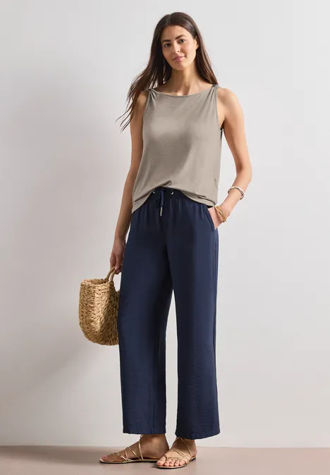 Frau in ärmellosem Top und weiten blauen Hosen, mit Strohtasche und Sandalen.