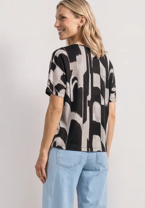 Eine Frau von hinten in einem schwarz-weißen, abstrakt gemusterten T-Shirt und hellblauen Jeans.