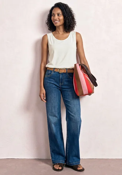 Vrouw in wit mouwloos top, wijde jeans en kleurrijke schoudertas voor een lichte achtergrond.