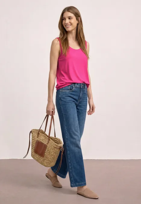 Femme en haut rose, jean bleu et chaussures beiges portant un sac en osier devant un fond neutre.