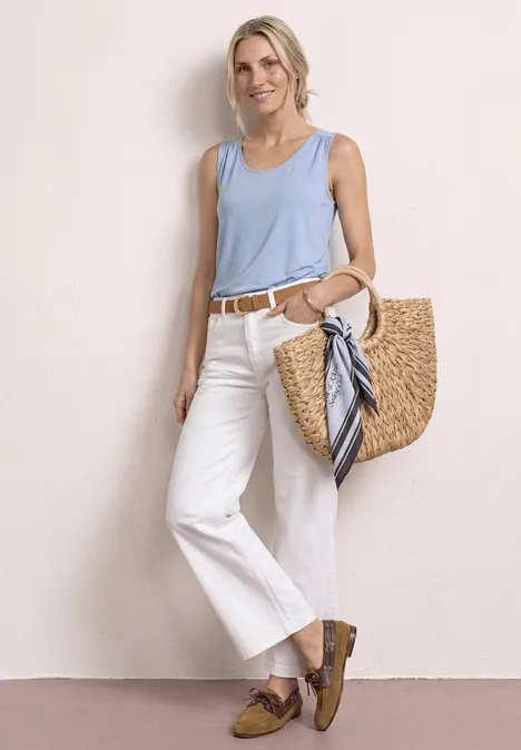 Top aus Jersey mit Elastiksaum cashmere blue Frau in hellblauem Top, weißer Hose, braunen Loafers und Korbtasche lehnt lässig an heller Wand.