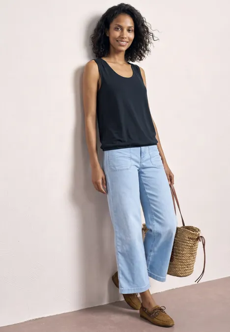 Top aus Jersey mit Elastiksaum shadowed navy Frau lehnt an heller Wand, trägt schwarzes Top, weite helle Jeans, braune Loafers und hält einen geflochtenen Korb.