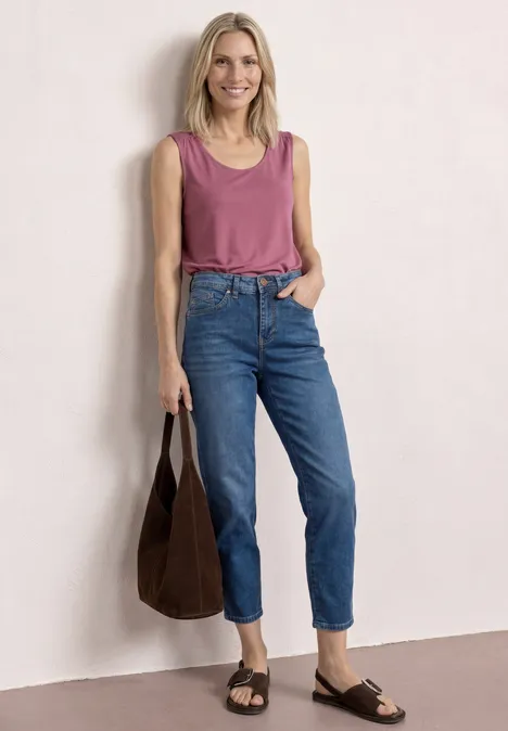 Vrouw in een roze top en blauwe jeans, met een donkere tas en sandalen, staand voor een lichte muur.