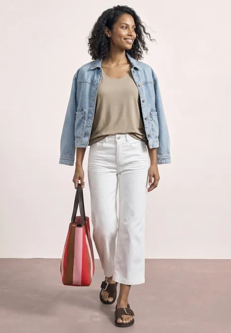 Vrouw in spijkerjasje, beige top, witte broek, sandalen en rode tas voor neutrale achtergrond.