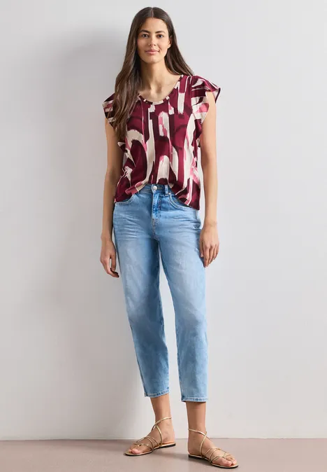Eine Frau trägt ein kurzärmeliges, gemustertes T-Shirt, blaue Jeans und Sandalen.