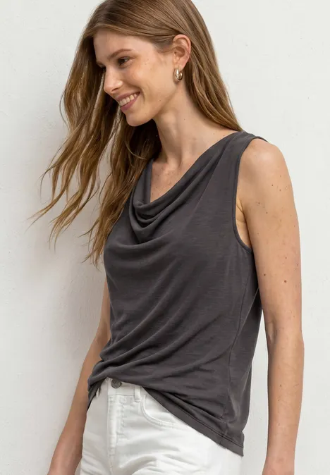 Top aspect soyeux avec décolleté cascade espresso brown Femme en haut sans manches gris foncé drapé et pantalon blanc, cheveux ondulés et boucles d’oreilles dorées.