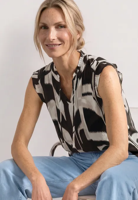Une personne assise porte un chemisier sans manches noir et blanc à motifs et un jean bleu clair.