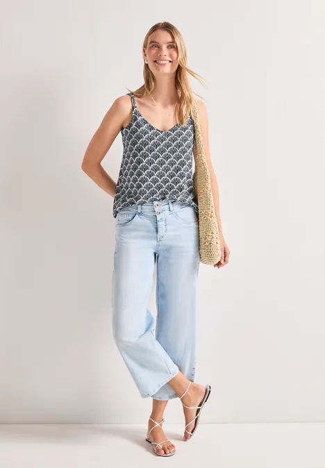 Frau trägt gemustertes Top, helle Jeans, weiße Sandalen und eine Strohtasche.