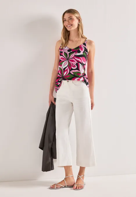 Frau in ärmelloser, bunt gemusterter Bluse, weißen Culottes, weißen Riemchensandalen, schwarze Jacke in der Hand.