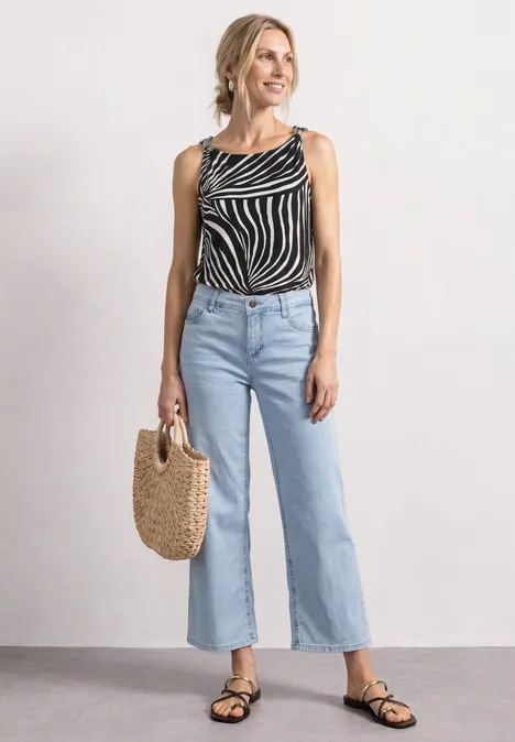 Frau mit Zebra-Print-Top, hellen Weitbein-Jeans, schwarzen Sandalen und Strohtasche.