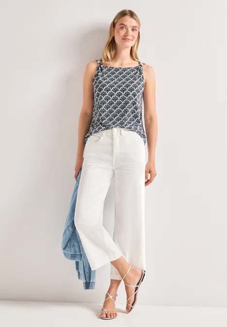 Frau im gemusterten Top, weißen Culottes und Sandalen, lässig mit hellblauer Jacke an der Seite.
