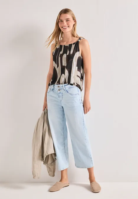 Femme portant un top imprimé, un jean large bleu clair et des mules plates, tenant une veste à la main.