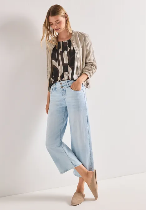 Femme portant une veste beige, un top à motifs, un jean large bleu clair et des mocassins beiges sur fond clair.