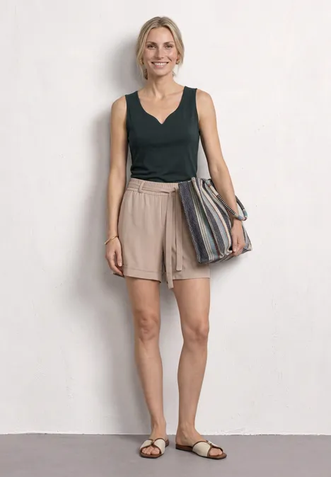 Basic Top mit Herzausschnitt deep bound green Eine schlanke Frau in dunkelgrünem Top, beigen Shorts und Sandalen hält eine gestreifte Tasche.