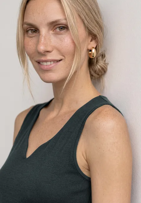 Basic Top mit Herzausschnitt deep bound green Eine blonde Frau mit Dutt, goldenen Creolen und ärmellosem Oberteil lächelt in die Kamera.