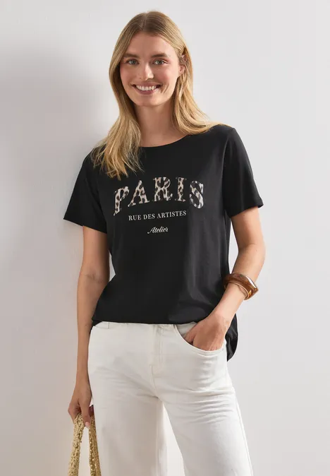 Frau in schwarzem 'PARIS'-T-Shirt und weißen Jeans, vor hellem Hintergrund, mit Tasche in der Hand.