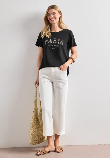Frau in schwarzem Paris-T-Shirt, weißer Culotte, flachen Sandalen und geflochtenem Korbtasche vor weißer Wand