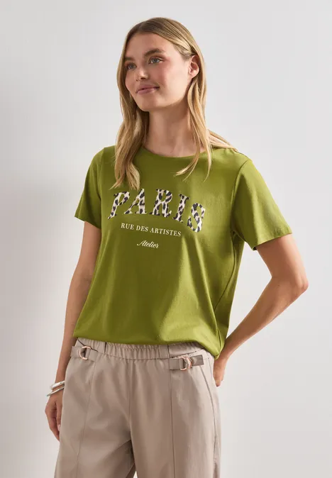 Person in grünem T-Shirt mit Print, beigen Hosen, seitlich vor weißem Hintergrund.