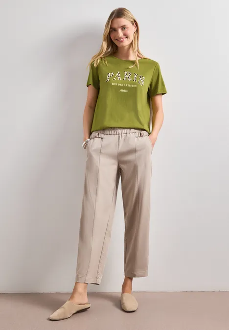 T-Shirt mit Wording Artwork shaded olive green Frau in grünem T-Shirt, beigen Stoffhosen und beigen Slippern vor weißer Wand.