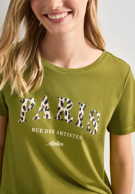 T-Shirt mit Wording Artwork shaded olive green Eine Frau trägt ein olivgrünes T-Shirt mit Leopardenmuster-Schriftzug «PARIS RUE DES ARTISTES Atelier».
