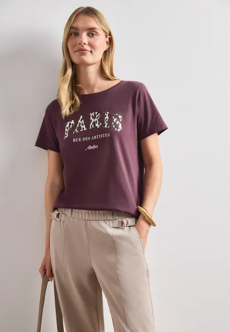 Frau trägt lila T-Shirt mit Schriftzug und helle Hose, steht und hält eine Tasche in der Hand.