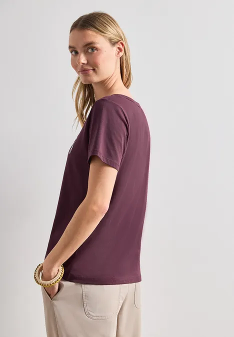 T-Shirt mit Wording Artwork rustic rouge Seitlich stehende Person in lila T-Shirt und beigen Hosen, das Gesicht unkenntlich gemacht.