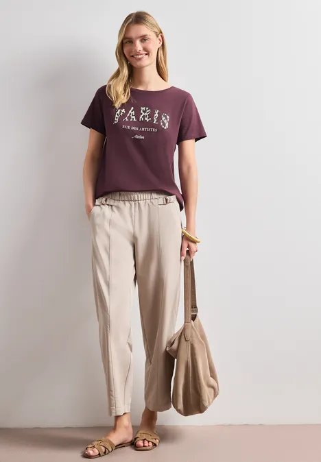 T-Shirt mit Wording Artwork rustic rouge Frau mit burgunderfarbenem T-Shirt mit 'PARIS', beigefarbenen Hosen und Tasche an der Seite.