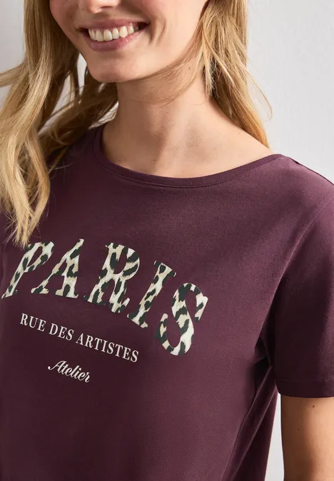 T-Shirt mit Wording Artwork rustic rouge Frau trägt Burgunderfarbenes T-Shirt mit Blumen-Print 'PARIS' und Schriftzug 'Rue des Artistes'.