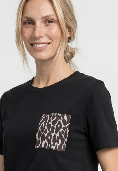 Eine Frau trägt ein schwarzes T-Shirt mit Leopardenmuster-Tasche vor hellem Hintergrund.