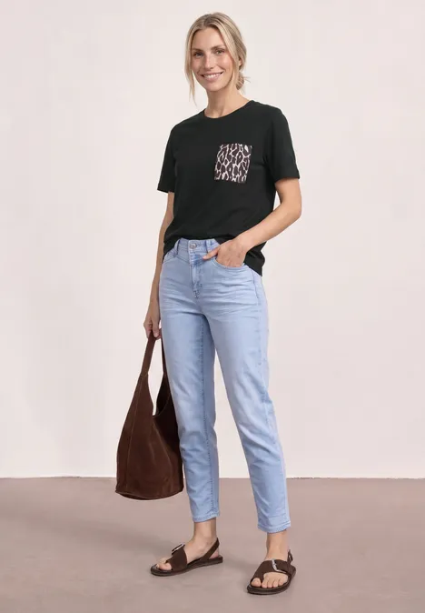 Frau trägt schwarzes T-Shirt mit gemusterter Brusttasche, hellblaue Jeans, schwarze Sandalen und eine braune Tasche.