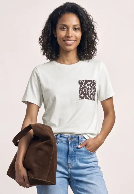 Eine Person trägt ein weißes T-Shirt mit gemustertem Brusttaschen-Detail und hellblaue Jeans, hält eine braune Jacke.