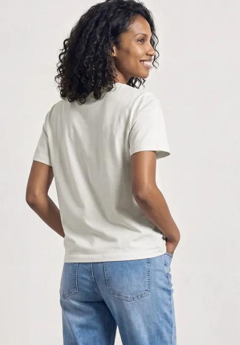 Eine Person steht mit dem Rücken zur Kamera, trägt weißes T-Shirt und blaue Jeans, lockige dunkle Haare.