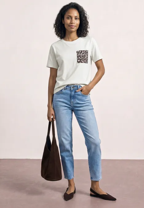 Frau in weißem T-Shirt mit gemusterter Brusttasche, blauen Jeans, schwarzen Ballerinas und brauner Tragetasche.