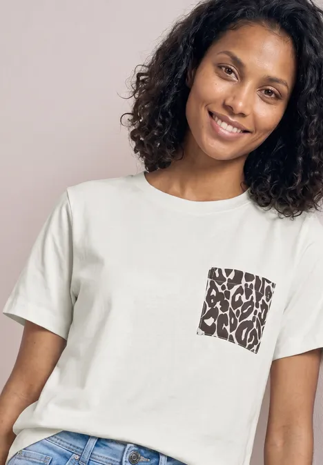 Eine Person trägt ein weißes T-Shirt mit einem kleinen quadratischen Brusttaschenmotiv und lockigem Haar.