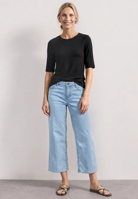 Vrouw in zwarte top, lichtblauwe cropped jeans en zwarte sandalen voor een lichte achtergrond.