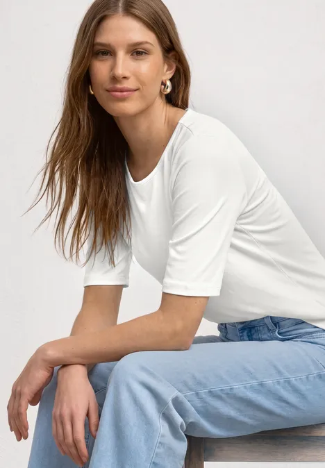 Een vrouw zit opzij, draagt een wit driekwart mouw T-shirt en lichtblauwe jeans.