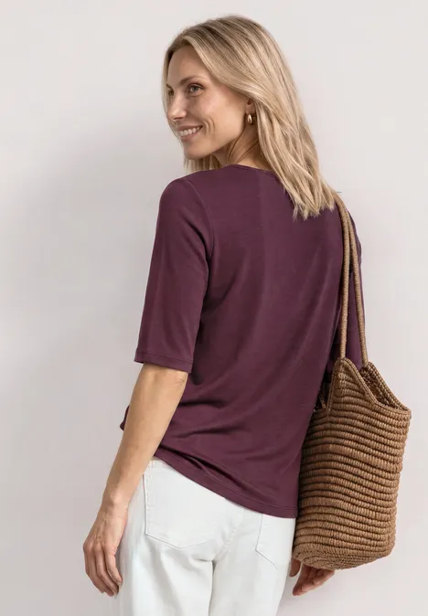 Basic Shirt met ronde hals in Unifarbe rustic rouge Vrouw met paarse T-shirt, witte broek en gevlochten tas, rug naar de camera en zijwaarts kijkend.