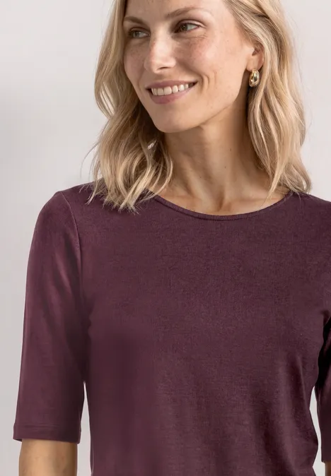 Basic Shirt met ronde hals in Unifarbe rustic rouge Een blonde vrouw glimlacht licht terwijl ze een bordeauxrood shirt met halve mouwen draagt voor een lichte achtergrond.